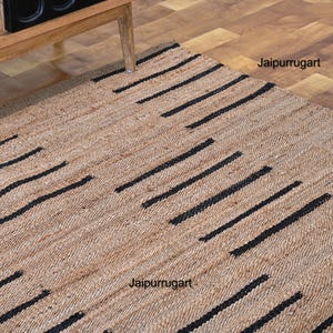 May include: A natural fiber rug with a geometric pattern of black stripes on a beige background. The rug is made of jute and is perfect for adding a touch of natural texture to any room. The rug is labeled "Jaipurruggart".