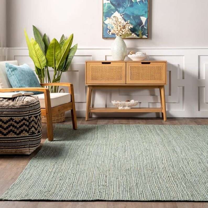 May include: A light blue jute rug with a woven texture. The rug is placed in a living room setting with a wooden chair, a side table, and a potted plant.