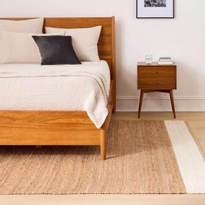 May include: A wooden bed frame with a light-colored textured blanket, two beige pillows, and one dark gray square pillow. Beside the bed sits a wooden nightstand with two drawers, a glass of water, and a small stack of books on top.