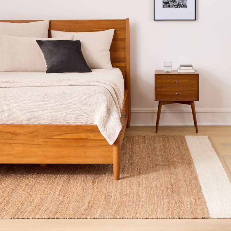 May include: A wooden bed frame with a light-colored textured blanket, two beige pillows, and one dark gray square pillow. Beside the bed sits a wooden nightstand with two drawers, a glass of water, and a small stack of books on top.