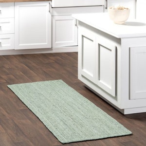 May include: A light green woven rug with a textured surface. The rug is placed on a wooden floor in a kitchen setting.