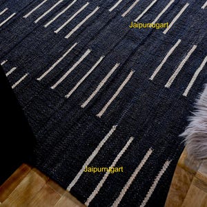 May include: A dark charcoal black area rug featuring a repeating pattern of short, vertical, off-white dashed lines arranged in staggered rows. The text Jaipurrugart is printed in yellow in two locations on the surface of the woven textile.