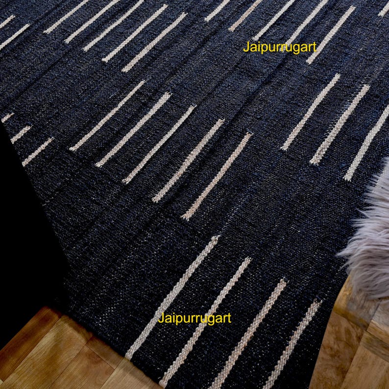 May include: A dark charcoal black area rug featuring a repeating pattern of short, vertical, off-white dashed lines arranged in staggered rows. The text Jaipurrugart is printed in yellow in two locations on the surface of the woven textile.