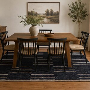 May include: A rectangular wooden dining table with four black spindle-back chairs featuring woven seats. The table is set with a large beige ceramic vase containing green branches and a wooden bowl. A dark blue rug with white stripes lies underneath.