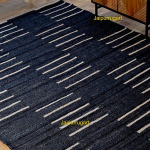 May include: A rectangular area rug featuring a dark navy blue woven texture with a repeating pattern of short, horizontal off-white stripes arranged in staggered rows. The text Jaipurrugart is visible in yellow on the surface of the rug.