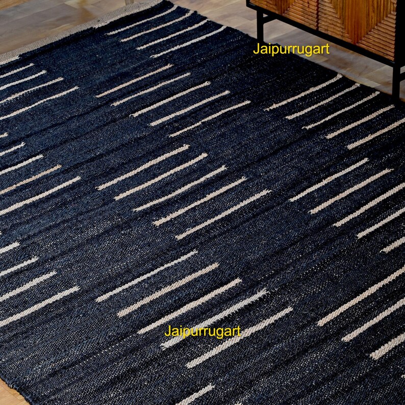 May include: A rectangular area rug featuring a dark navy blue woven texture with a repeating pattern of short, horizontal off-white stripes arranged in staggered rows. The text Jaipurrugart is visible in yellow on the surface of the rug.