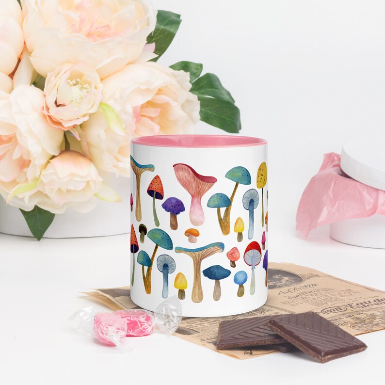 May include: A white ceramic mug with a pink rim features a colorful watercolor illustration of various mushrooms. The mug is filled with a light brown liquid.