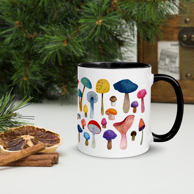 May include: White ceramic mug with a black rim and handle. The mug features a colorful watercolor illustration of various mushrooms.