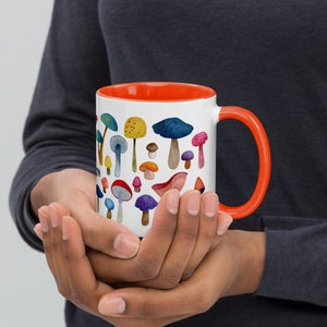 May include: A white ceramic mug with an orange rim and handle. The mug features a colorful watercolor illustration of various mushrooms.