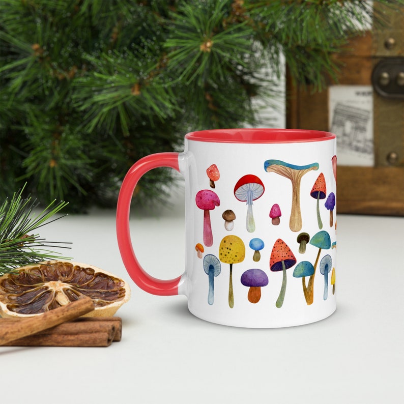May include: A white ceramic mug with a red rim features a colorful watercolor illustration of various mushrooms. The mug is perfect for enjoying a hot beverage.