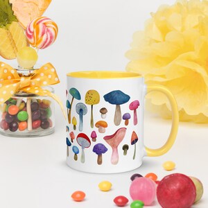 May include: A white ceramic mug with a yellow rim features a colorful watercolor illustration of various mushrooms. The mug is filled with a white liquid and is sitting on a white surface.