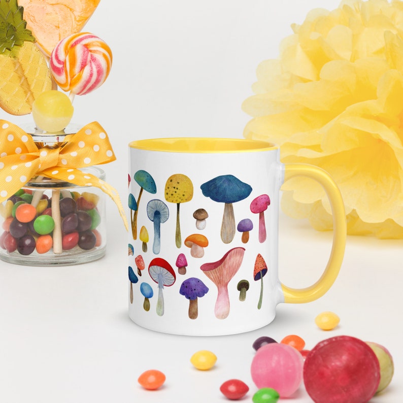 May include: A white ceramic mug with a yellow rim features a colorful watercolor illustration of various mushrooms. The mug is filled with a white liquid and is sitting on a white surface.
