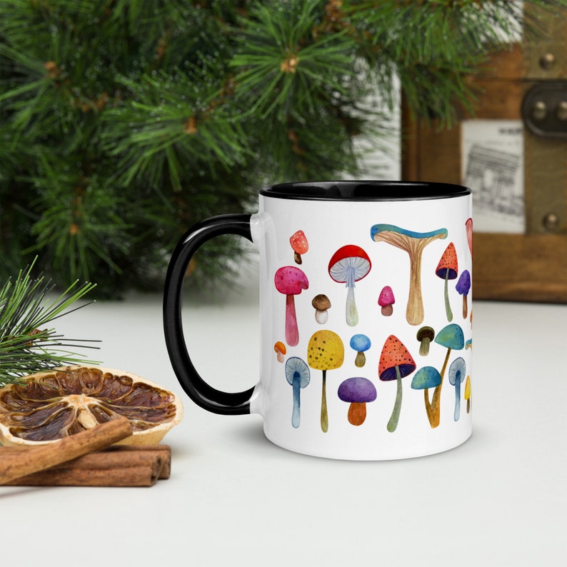 May include: White ceramic mug with a black rim and handle. The mug features a colorful illustration of various mushrooms with different shapes and colors.