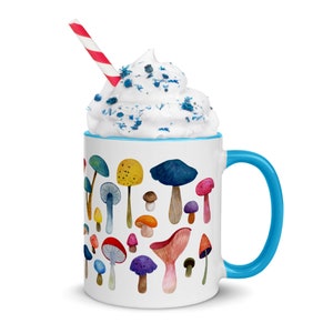 May include: A white ceramic mug with a blue rim and handle. The mug is filled with whipped cream and blue sprinkles. The mug has a design of colorful mushrooms on a white background. A red and white striped straw is sticking out of the whipped cream.