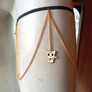 May include: A gold chain leg garter with a small white cat charm. The cat charm has a crescent moon on its forehead and a red heart on its chest.