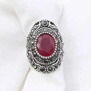 Peut inclure: Bague argentée ornée d'une pierre précieuse ovale rouge foncé au centre. La bague est entourée de petites pierres grises foncées et de détails ornés. Posée sur un fond de tissu blanc.