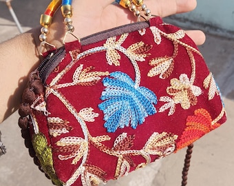 Könnte beinhalten: Eine kleine, rote Samt-Handtasche mit einem floralen Stickmuster. Die Tasche hat einen oberen Griff aus bunter geflochtener Schnur und einen langen Schultergurt.