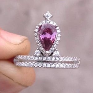 以下が含まれることがあります： 梨型のピンクの宝石を冠型のセッティングで飾ったリング。バンドには小さなダイヤモンドが並んでいます。