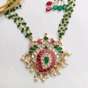 May include: A gold necklace with a large, ornate pendant featuring a peacock design. The pendant is decorated with red, green, and white gemstones. The necklace is strung with green beads and has a gold tassel.