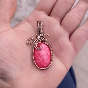Peut inclure: Un pendentif artisanal avec une pierre rose de forme ovale, sertie dans du fil de cuivre. Le travail du fil comprend des volutes décoratives et un dessus tissé, créant un bijou unique et artistique. Le pendentif mesure environ 3,8 cm de long.