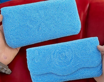 May include: Two light blue beaded clutch purses with floral designs. The purses are rectangular with rounded edges and a flap closure. The beadwork creates intricate patterns, and the purses are held against a red background.