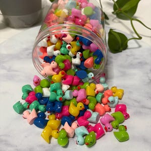 May include: A clear plastic jar tipped over, spilling out a collection of colorful miniature duck figurines. The ducks are in various shades of pink, blue, green, yellow, and white. Some ducks are upright, while others are scattered around the jar.