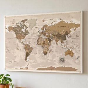 Può includere: Una mappa del mondo incorniciata in tonalità marroni e beige, montata su una parete bianca. La mappa presenta informazioni geografiche dettagliate e una rosa dei venti. La cornice è bianca e la mappa è sopra un tavolo di legno.