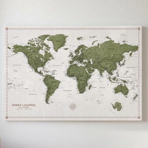 May include: A rectangular world map wall art piece featuring olive green landmasses against a light cream background. The map includes a decorative border, a compass rose, and the printed text EMILY & OLIVER OUR TRAVELS in the bottom left corner.