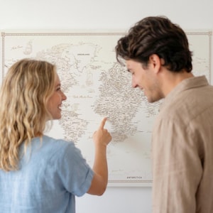 Può includere: Una mappa del mondo incorniciata in tonalità neutre è appesa a una parete bianca. Una coppia guarda la mappa, con una persona che indica un luogo. La mappa presenta informazioni geografiche dettagliate.