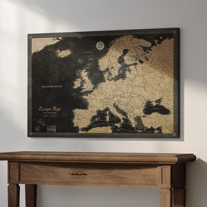 Könnte beinhalten: Eine rechteckige Wandkarte von Europa mit schwarzem Hintergrund und goldfarbenen Landmassen und Küstenlinien. Die Karte enthält den Text Europe Map, Atlantic Ocean und eine Windrose. Sie ist in einem dünnen schwarzen Rahmen an der Wand befestigt.