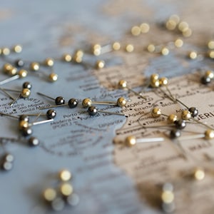 Peut inclure: Gros plan d'une carte du monde avec des punaises dorées et argentées marquant différents endroits. La carte est partiellement masquée par les punaises, mais les mots "MAROC" et "NIGÉRIA" sont visibles.