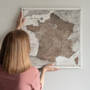 Könnte beinhalten: Eine Person hängt eine Vintage-Karte von Frankreich an eine weiße Wand. Die Karte ist auf Leinwand gedruckt und zeigt eine detaillierte Umrisskarte der Regionen des Landes. Die Karte trägt den Titel "La France" und enthält eine Kompassrose.