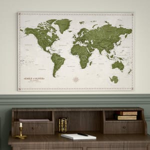 Peut inclure: Une carte du monde encadrée avec un design en relief détaillé dans des tons verts, montée sur un fond crème. La carte est étiquetée avec des noms de lieux et présente une bordure décorative. Le texte "EMILY & OLIVER" est visible. La carte est affichée au-dessus d'un bureau en bois.