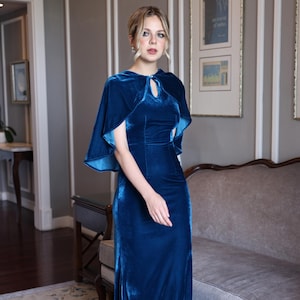 Könnte beinhalten: Ein langes, dunkelblaues Samtkleid mit einem Cape. Das Kleid hat einen Schlüssellochausschnitt und einen hohen Schlitz. Das Cape hat ein fließendes Design. Das Kleid ist bodenlang und figurbetont. Das Bild ist in Innenräumen aufgenommen.