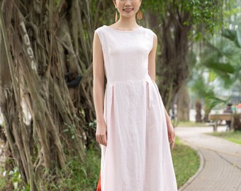 May include: A woman wearing a long, light pink linen dress with a high neckline and a side slit. She is standing on a brick pathway in a park setting.