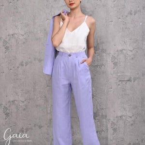 Full-length shot of a female model holding her blazer over her shoulder, showcasing the high-rise waistband with a seamless button and zipper fly closure on the lilac linen trousers.