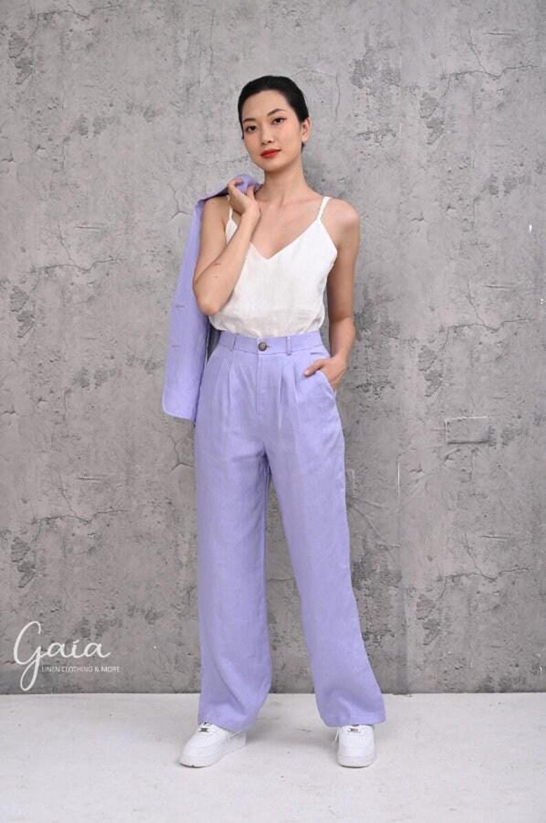 Full-length shot of a female model holding her blazer over her shoulder, showcasing the high-rise waistband with a seamless button and zipper fly closure on the lilac linen trousers.