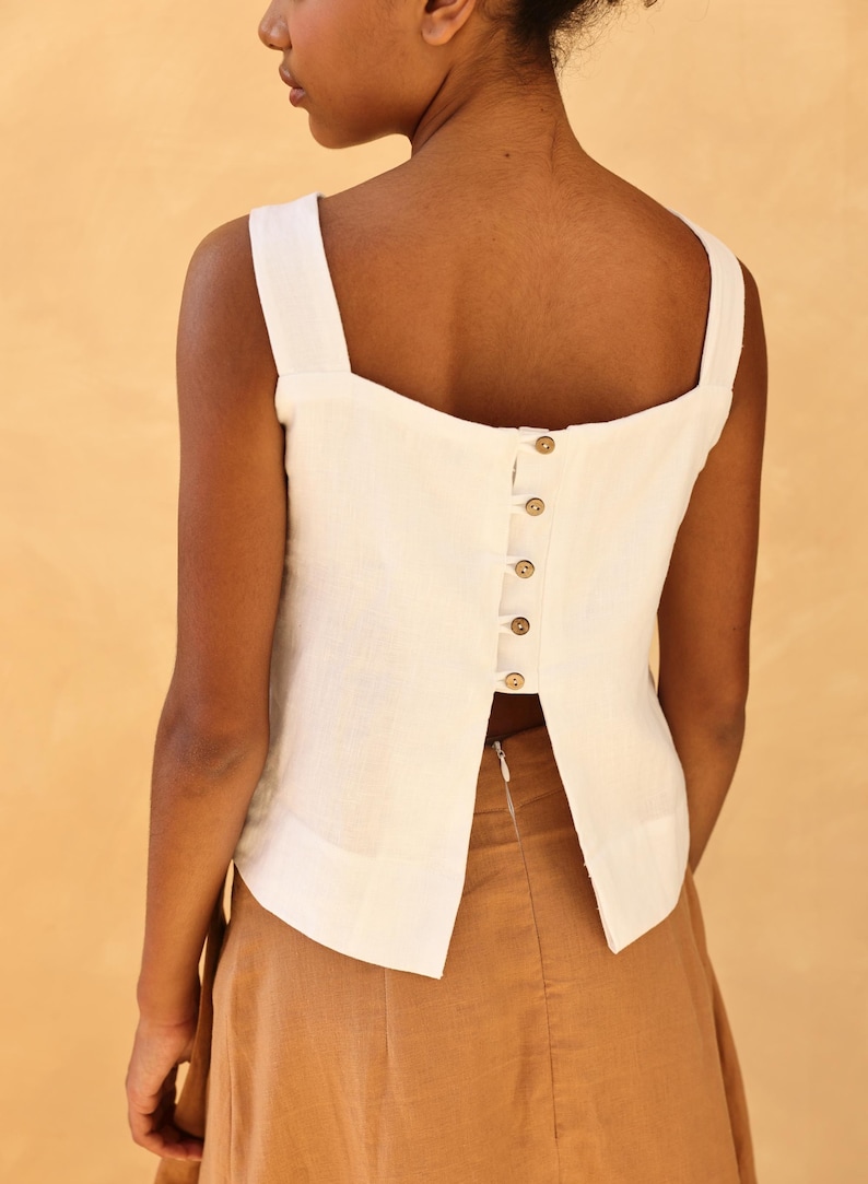 A medium rear close-up shot focusing on the back bodice layout of a model wearing a white linen tank top. The image showcases the square back neckline, wide shoulder straps, and a central back button closure with a dramatic split-back detail opening below the waist.