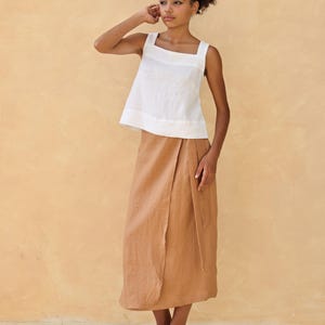 A full-length three-quarter portrait of the model posing dynamically with one hand raised to her shoulder. The soft warm lighting defines the clean French seams and highlights the effortless, breezy style of the two-piece linen outfit.