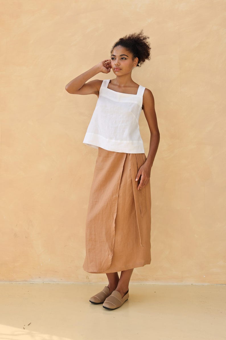 A full-length three-quarter portrait of the model posing dynamically with one hand raised to her shoulder. The soft warm lighting defines the clean French seams and highlights the effortless, breezy style of the two-piece linen outfit.
