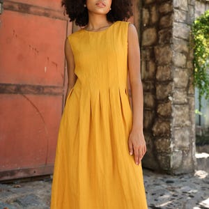 May include: A sleeveless, mustard yellow dress with a pleated bodice and a flowing skirt. The dress is made of a textured fabric and has a knee-length hemline. The model is standing in front of a red door and stone wall.