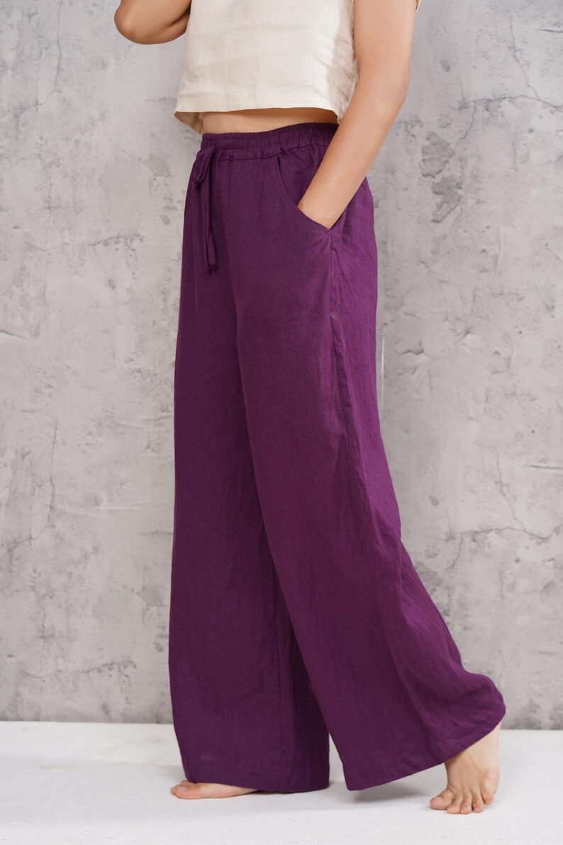 Three-quarter front full-body view of the model posing with her hand in her pocket against the studio wall, highlighting the casual chic elegance of the purple linen ensemble.