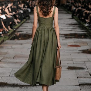 Custom olive linen wedding guest dress in minimalist A-line, back view, sleeveless with wide straps, walking a stone runway.