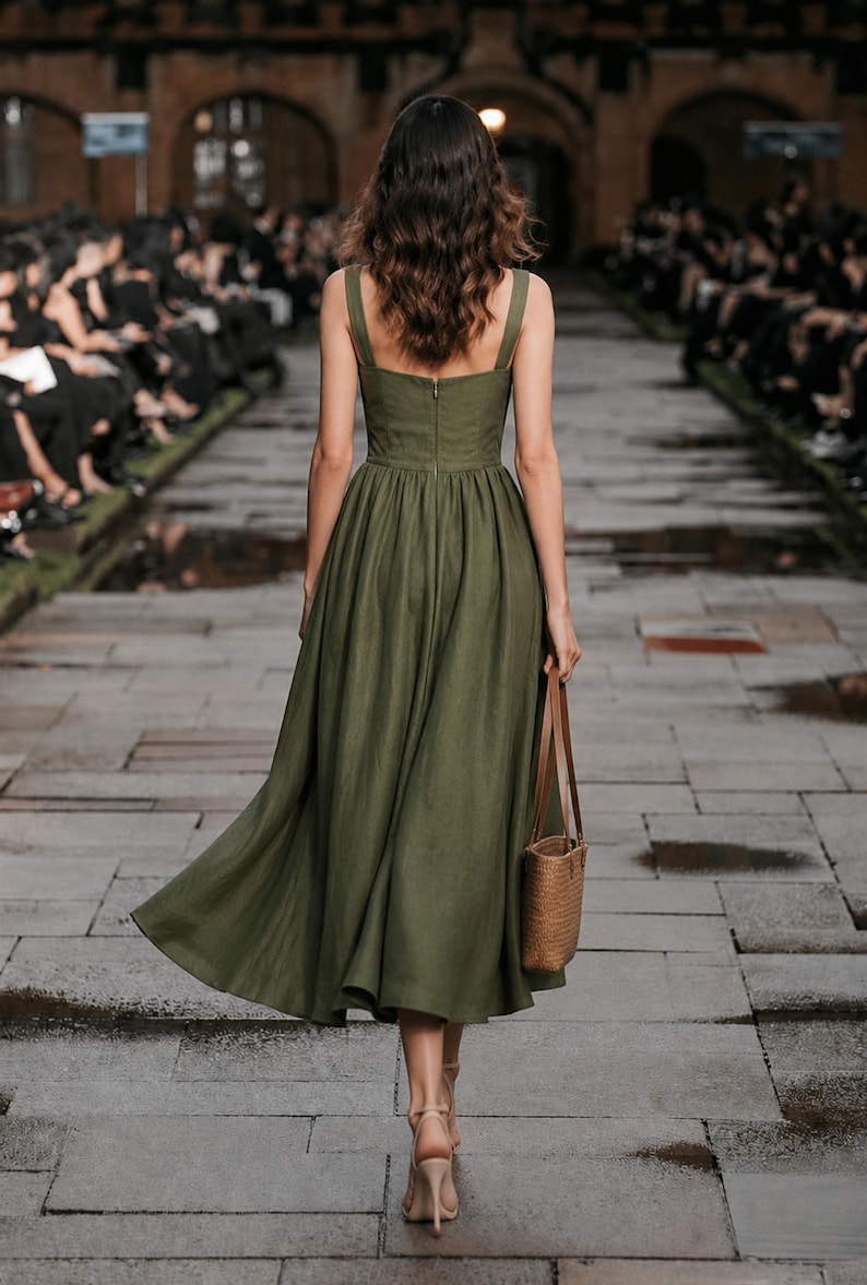 Custom olive linen wedding guest dress in minimalist A-line, back view, sleeveless with wide straps, walking a stone runway.