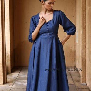 Medium front view from the thighs up of a woman looking down with a relaxed posture, highlighting the functional side pockets, gathered waist, and rich texture of a blue linen summer dress.