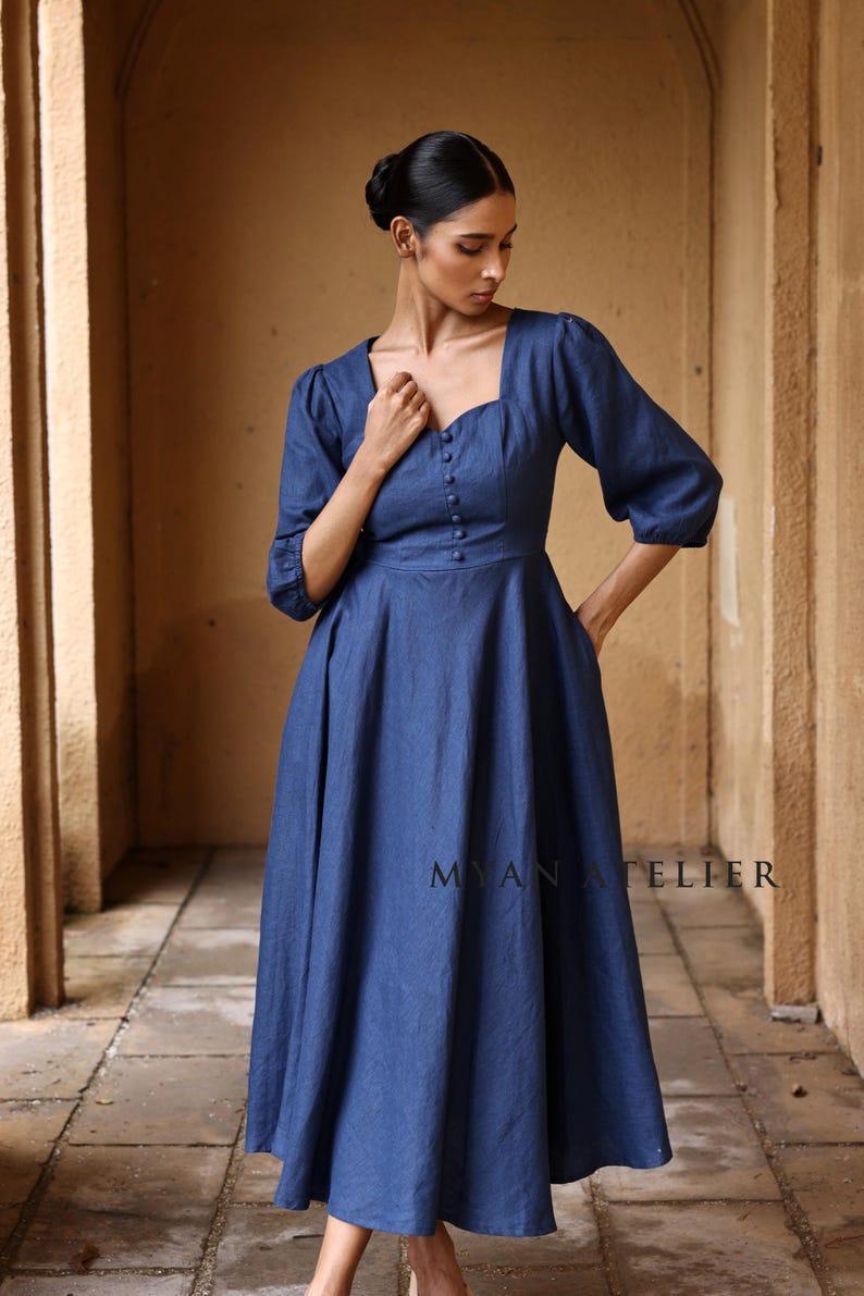 Medium front view from the thighs up of a woman looking down with a relaxed posture, highlighting the functional side pockets, gathered waist, and rich texture of a blue linen summer dress.