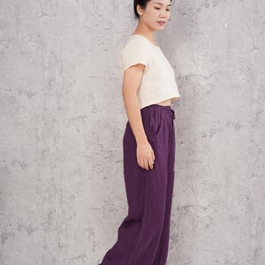 Side-profile full-body view of the model standing in the studio, showcasing the clean vertical line, generous length, and elegant loose flow of the purple wide-leg pants.