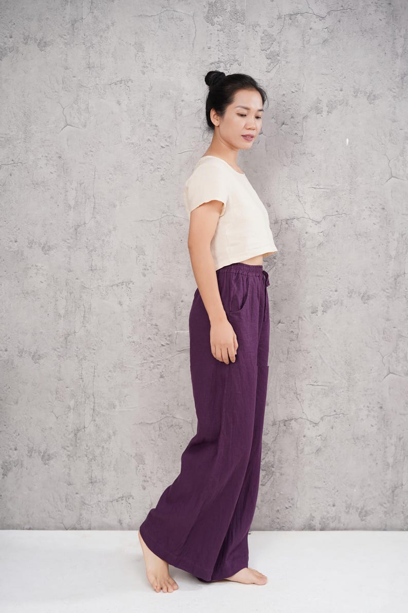 Side-profile full-body view of the model standing in the studio, showcasing the clean vertical line, generous length, and elegant loose flow of the purple wide-leg pants.