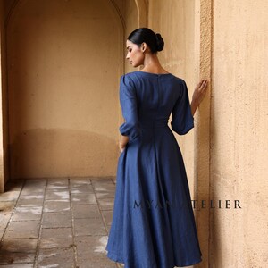 Full-length back and side three-quarter view of a woman looking away down a sunlit architectural hallway, demonstrating the modest high back design and elegant A-line silhouette of a blue linen dress.