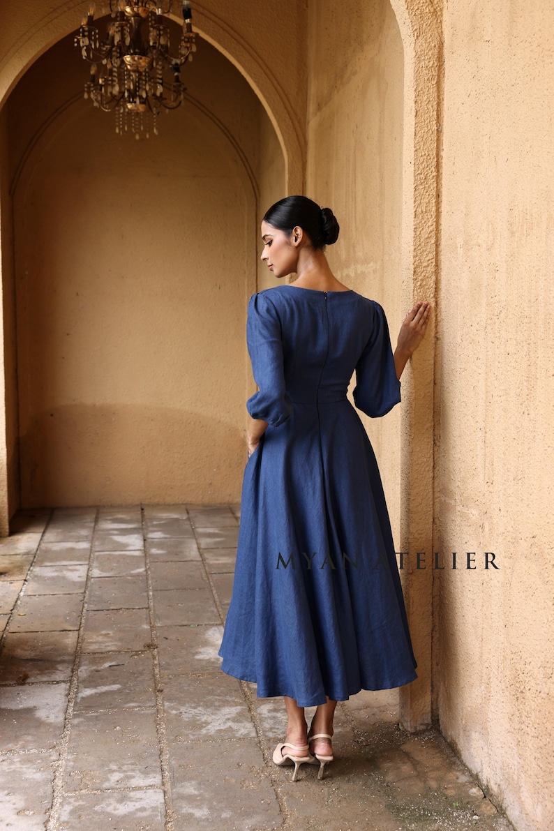 Full-length back and side three-quarter view of a woman looking away down a sunlit architectural hallway, demonstrating the modest high back design and elegant A-line silhouette of a blue linen dress.